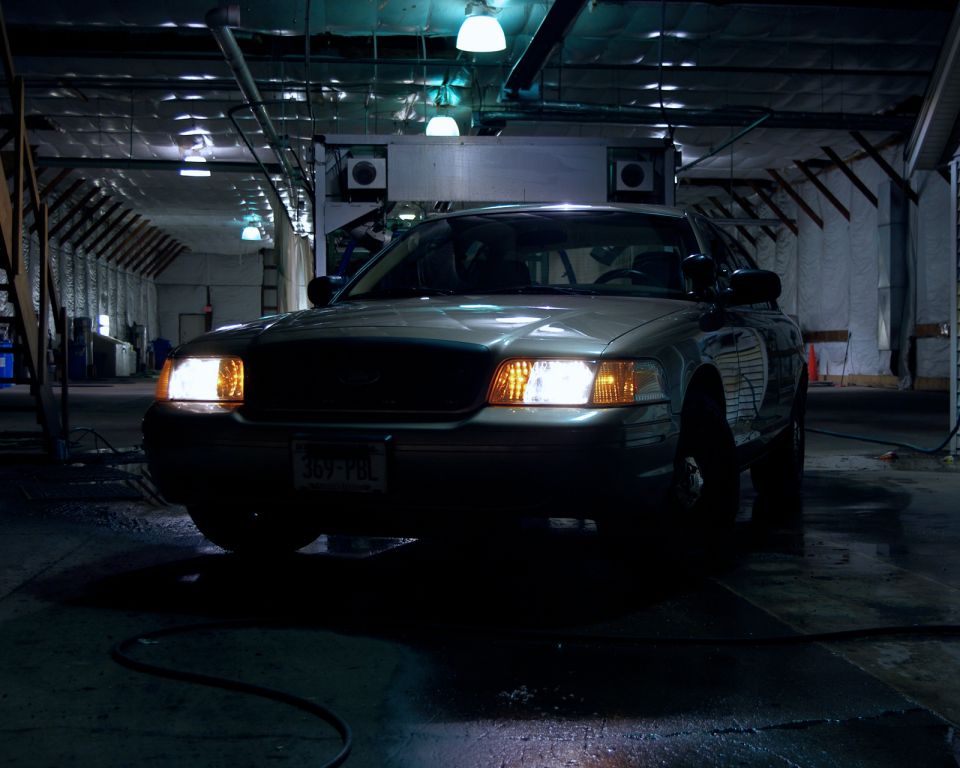 Tan Crown Victoria sedan with headlights on inside a dark warehouse, plate 369-PBL