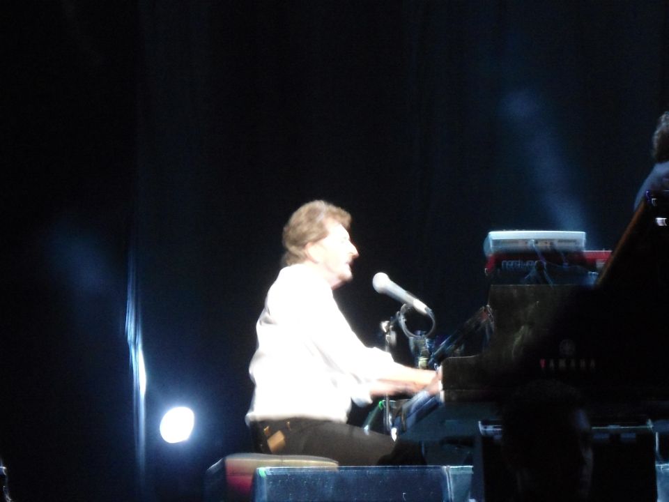 Man in white shirt playing a black grand piano on stage with a microphone