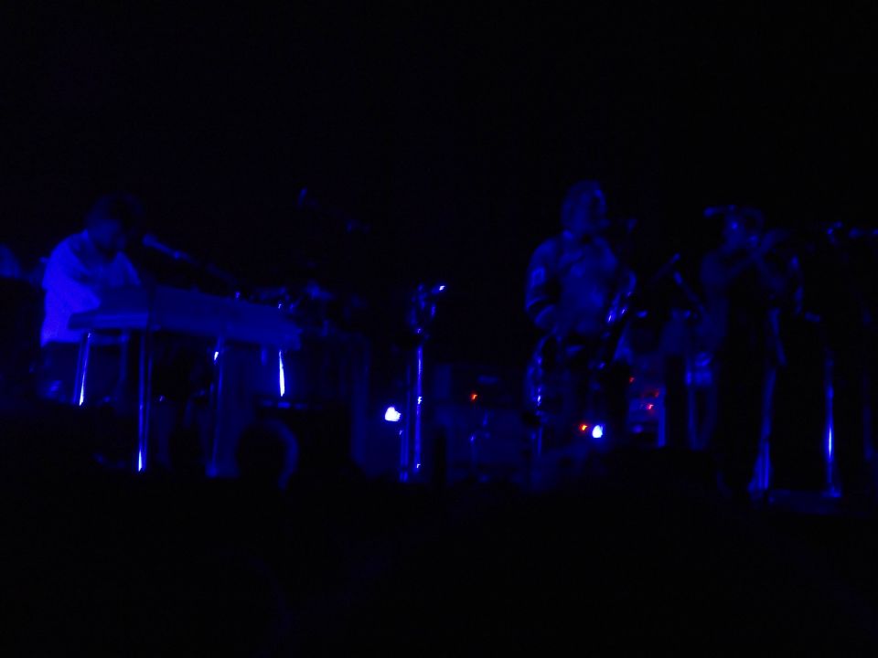Musicians playing keyboards and drums on a dark stage illuminated by blue lights