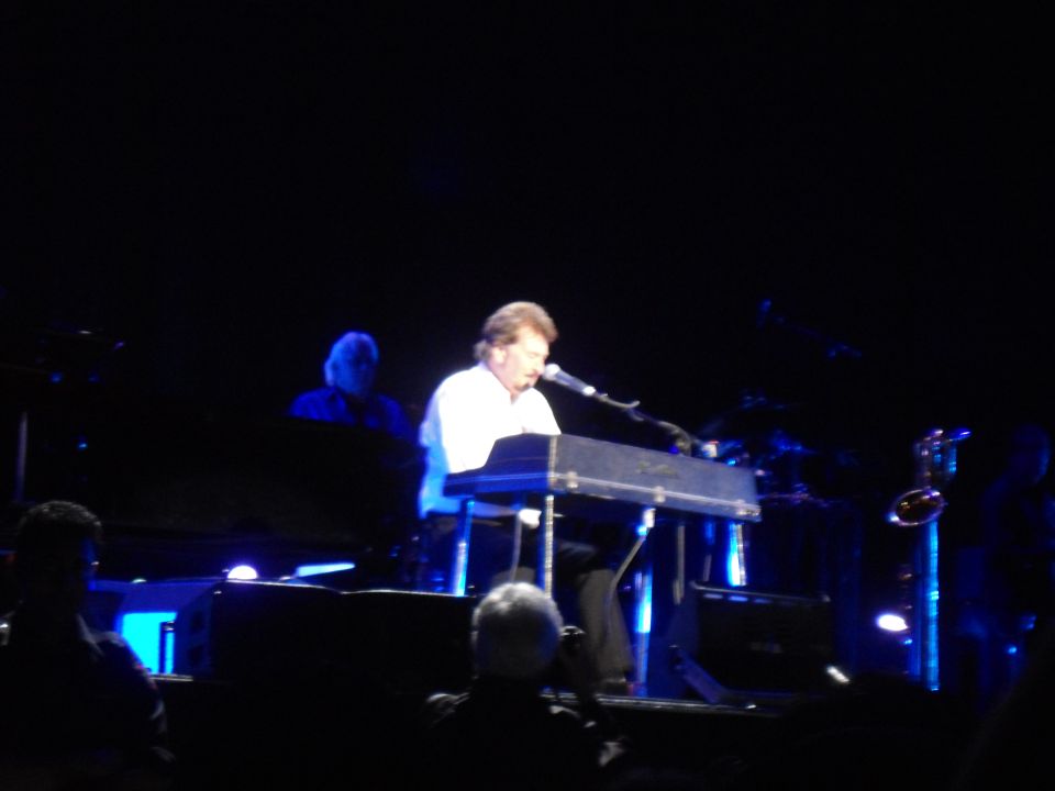 Man in white shirt plays keyboard on stage with blue lights and other musicians in background