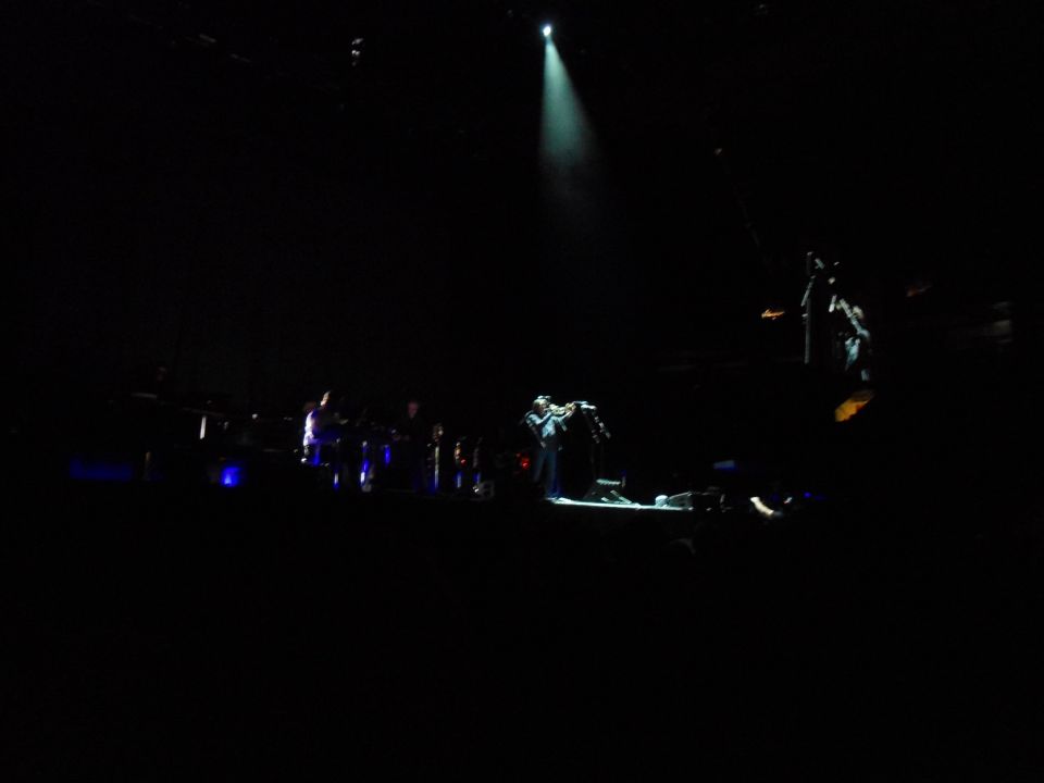 Musician plays violin on stage under spotlight with band behind