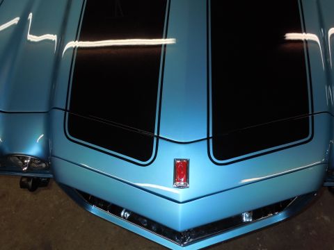 1972 Chevrolet Camaro - Close-up of a blue car's front end, showing racing stripes and a red emblem.