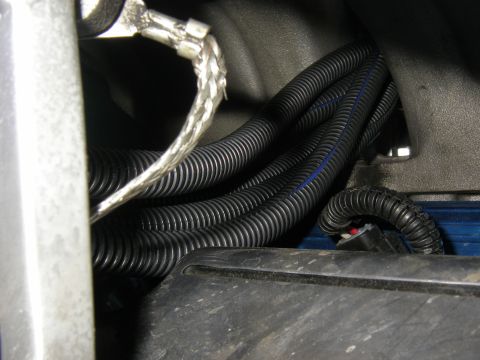 1990 Ford Bronco - Tight engine-bay shot of black split-loom convoluted wiring conduit and a braided-sleeve ground strap routed past a bare metal bracket. A s…