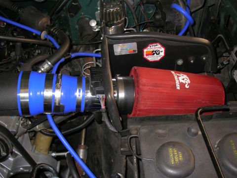 1990 Ford Bronco - Engine-bay view of an aftermarket cold-air intake: a red K&N conical filter with a black heat-shield bracket feeds a polished aluminum tube…