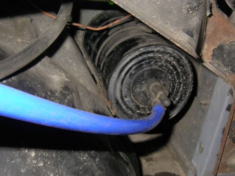 1990 Ford Bronco - Grimy round steel vacuum reservoir canister tucked behind the inner fender, with a blue silicone vacuum hose routed onto one of its ports a…