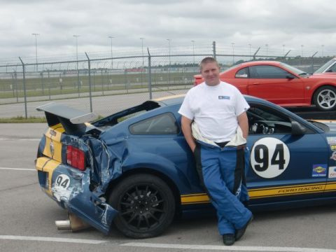 2007 Ford Mustang - A man stands beside a damaged blue 2007 Ford Mustang race car on a track. A red Mustang is parked behind it.