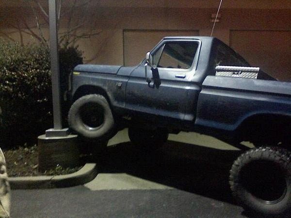 Lifted dark blue Ford pickup at night with front tire up on a parking curb, toolbox in bed