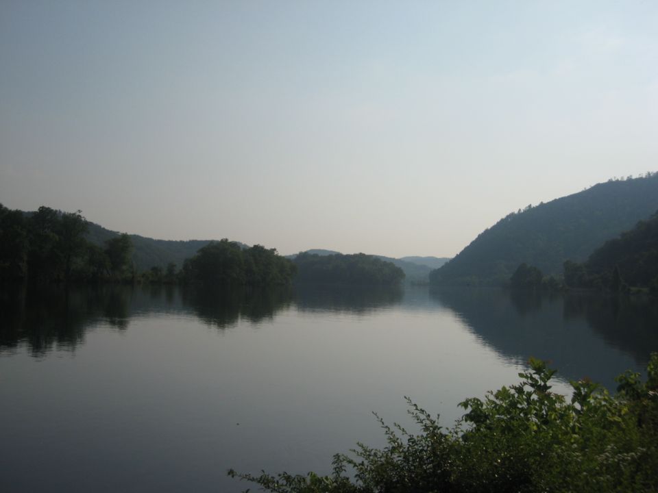 Calm lake reflects tree-covered hills under a hazy sky