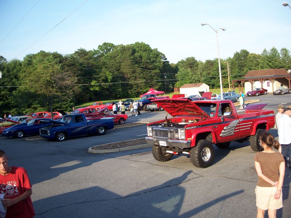 Red pickup truck with hood open, parked at an outdoor car show with other vehicles