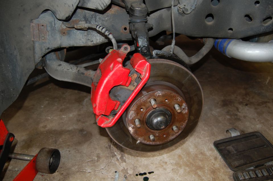red brake caliper over a rusty rotor on a car's suspension. A blue and silver pipe is visible in the background