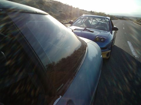 1995 Ford Probe - Two cars, one a 1995 Ford Probe with a roll cage, are parked on a road with a scenic background.