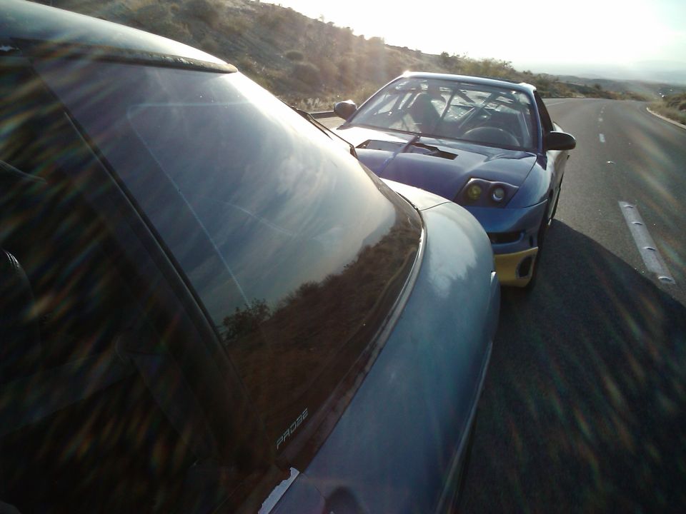 Blue 1995 Ford Probe with roll cage visible, parked on a road next to another car