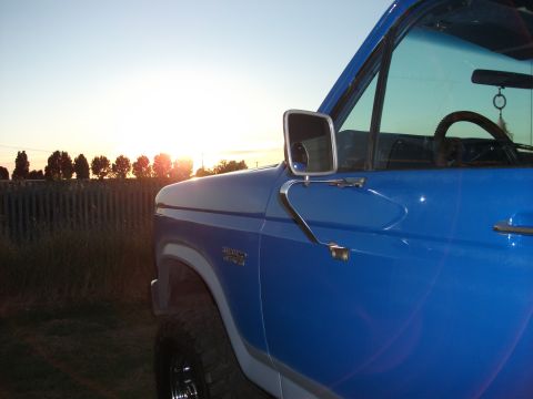 1982 Ford Bronco - A blue 1982 Ford Bronco is seen from the side at sunset, with a wooden fence and trees in the distance.