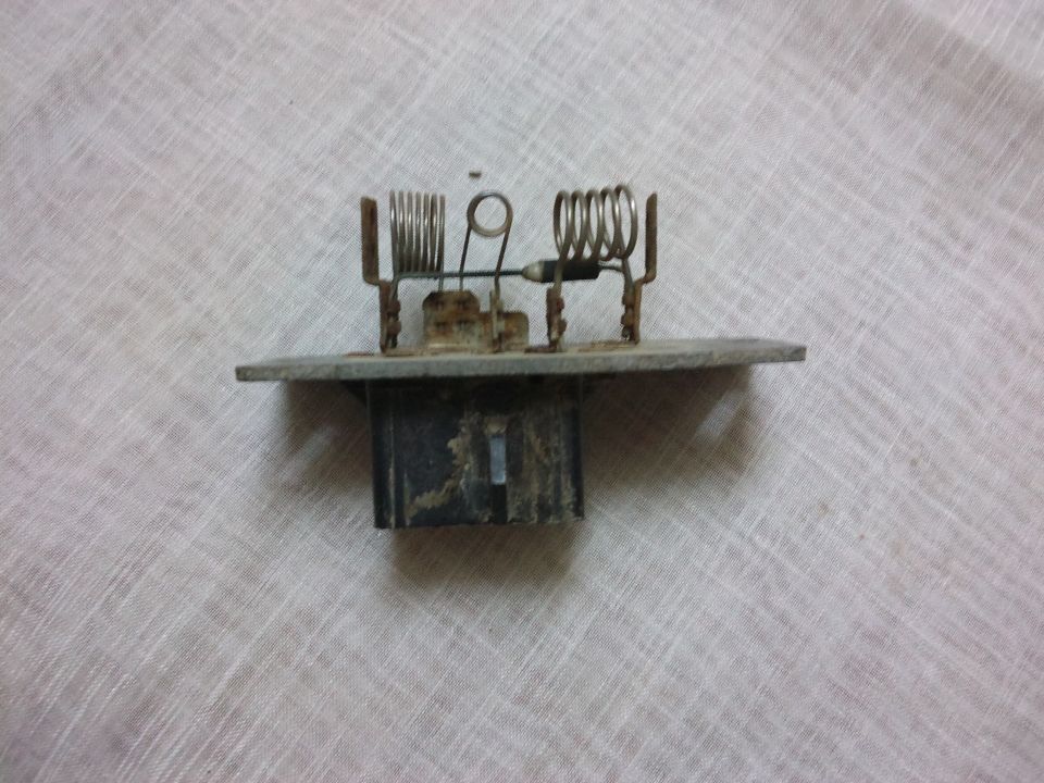 Blower motor resistor with metal coils and a black plastic connector, resting on a textured surface
