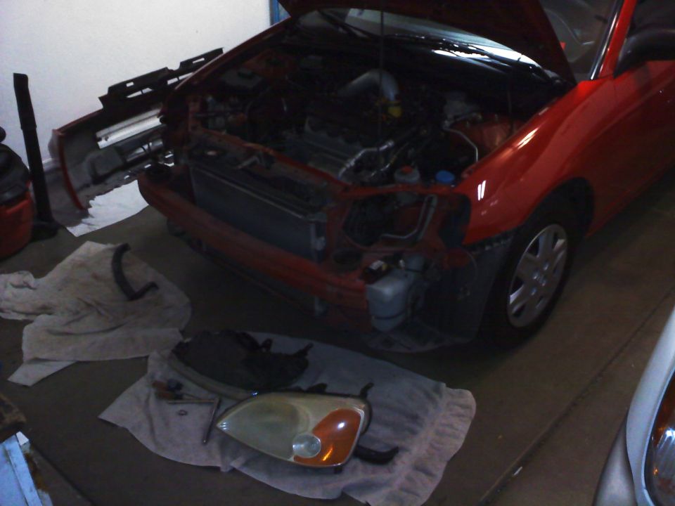 red car's front end is taken apart, with the bumper and headlights removed and placed on the ground