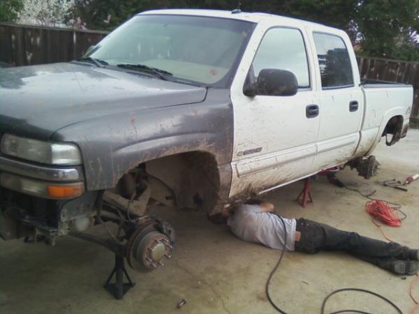 White/gray Silverado pickup on jack stands with front wheel off, person working underneath