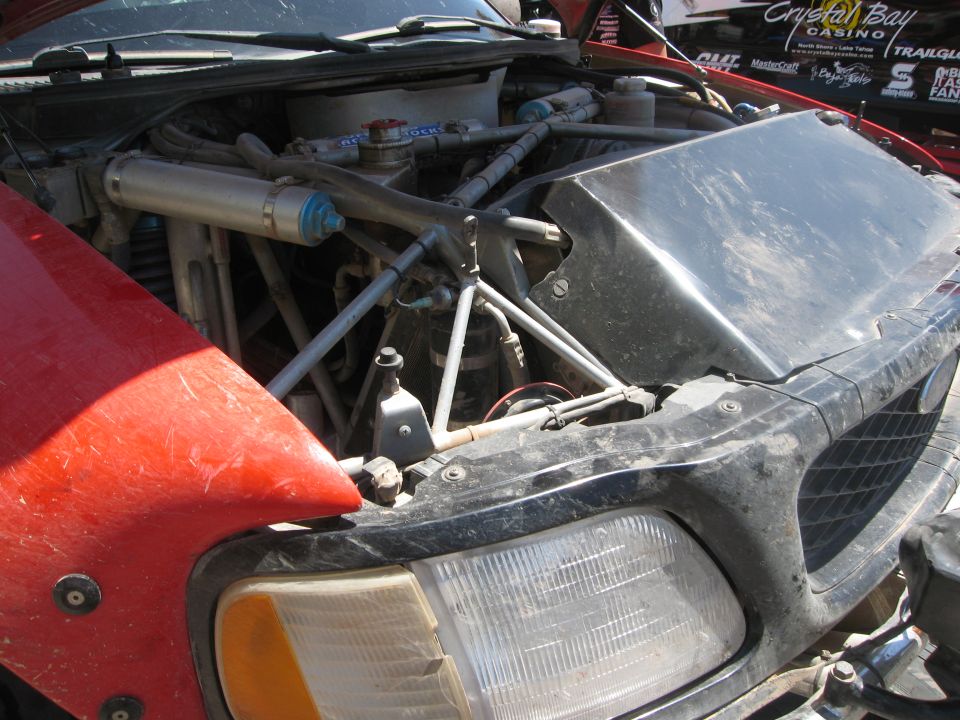 Engine bay of a red vehicle with custom modifications. Shows engine components, suspension parts, and some racing decals in the background
