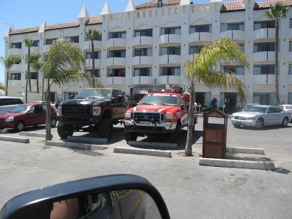 Two lifted pickup trucks, one black with Rockstar graphics and one red, parked in a lot. A silver sedan is parked to the right