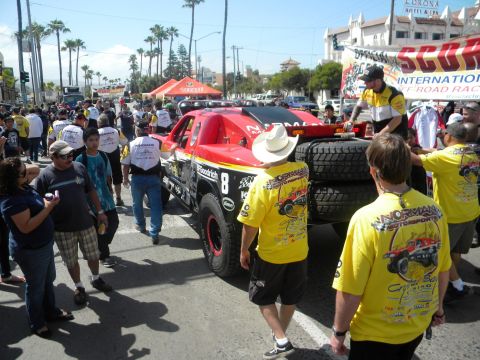 1986 Ford Bronco - A crowd gathers around a red off-road race truck with spare tires mounted on the rear, during an event.