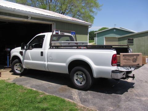2008 Ford F-250 - A white Ford F-250 pickup truck is parked in front of a garage, its bed loaded with a toolbox and several boxes.