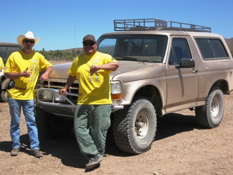 1986 Ford Bronco - A tan Ford Bronco is parked on a dirt lot with two men standing next to it under a clear blue sky.