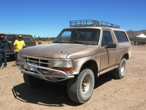 1986 Ford Bronco - A tan Ford Bronco with a roof rack and aftermarket bumper is parked on a dirt lot with two people nearby.