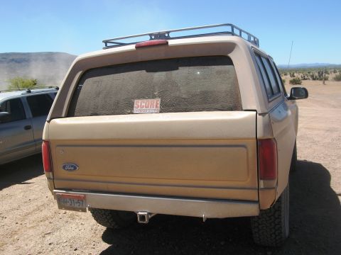 1986 Ford Bronco - Rear view of a tan Ford Bronco, a SCORE sticker and a roof rack, parked in a desert landscape.