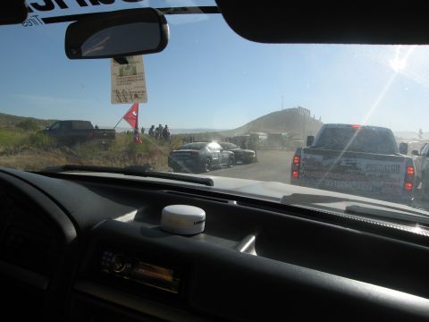 1986 Ford Bronco - A view from inside a vehicle shows a line of cars and trucks on a dusty road with a hill in the background.
