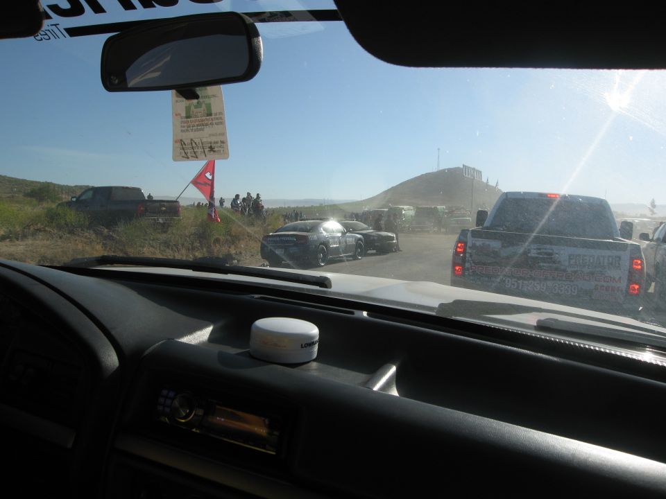 Pickup truck with "PREDATOR OIL" on the back, driving on a dirt road with other cars and people