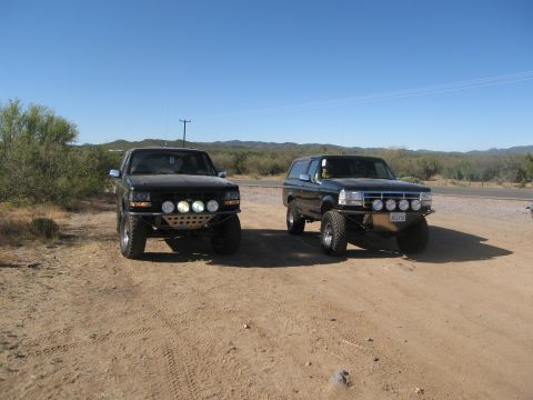 1986 Ford Bronco - Two Ford Broncos are parked side-by-side on a dusty terrain under a clear blue sky.