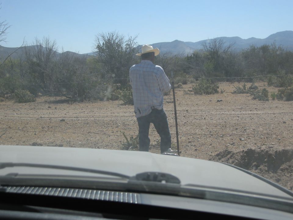 Man in cowboy hat and plaid shirt stands in a dry, brushy area with mountains in the background