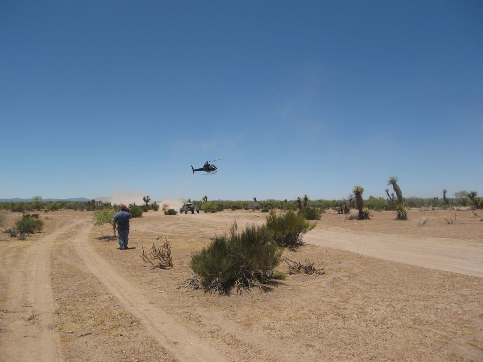 Desert scene with a helicopter flying overhead, a person walking on a dirt road, and two off-road vehicles in the distance
