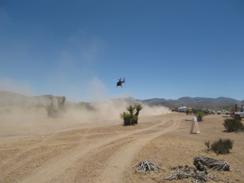 1986 Ford Bronco - A helicopter observes a dusty off-road race event in a desert landscape with spectators and vehicles.