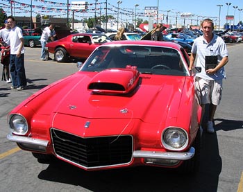 1991 Ford Bronco - A man stands next to a bright red muscle car from the 1970s, likely a Chevrolet Camaro, at an outdoor car show.