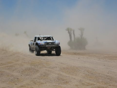 1986 Ford Bronco - A modified truck races through a dusty desert landscape under a clear blue sky.