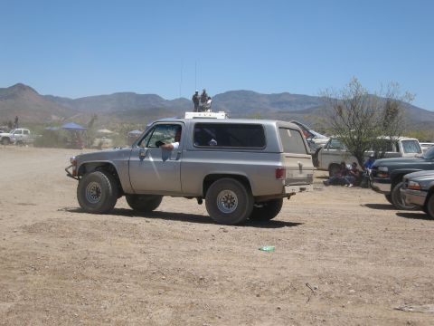 1986 Ford Bronco - A silver SUV with off-road tires is parked in a dusty outdoor area with other vehicles and people.
