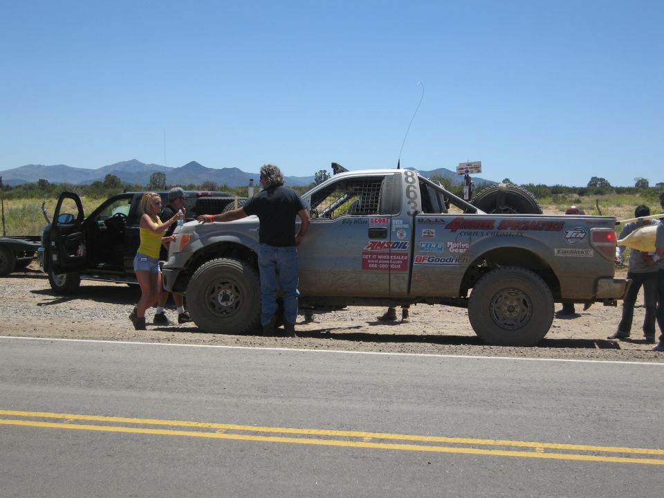 Off-road truck with racing decals and a spare tire on back, people around it. Another truck and trailer nearby