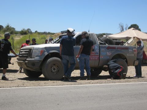 1986 Ford Bronco - A group of people attend to an off-road pickup truck with numerous racing decals on its side.