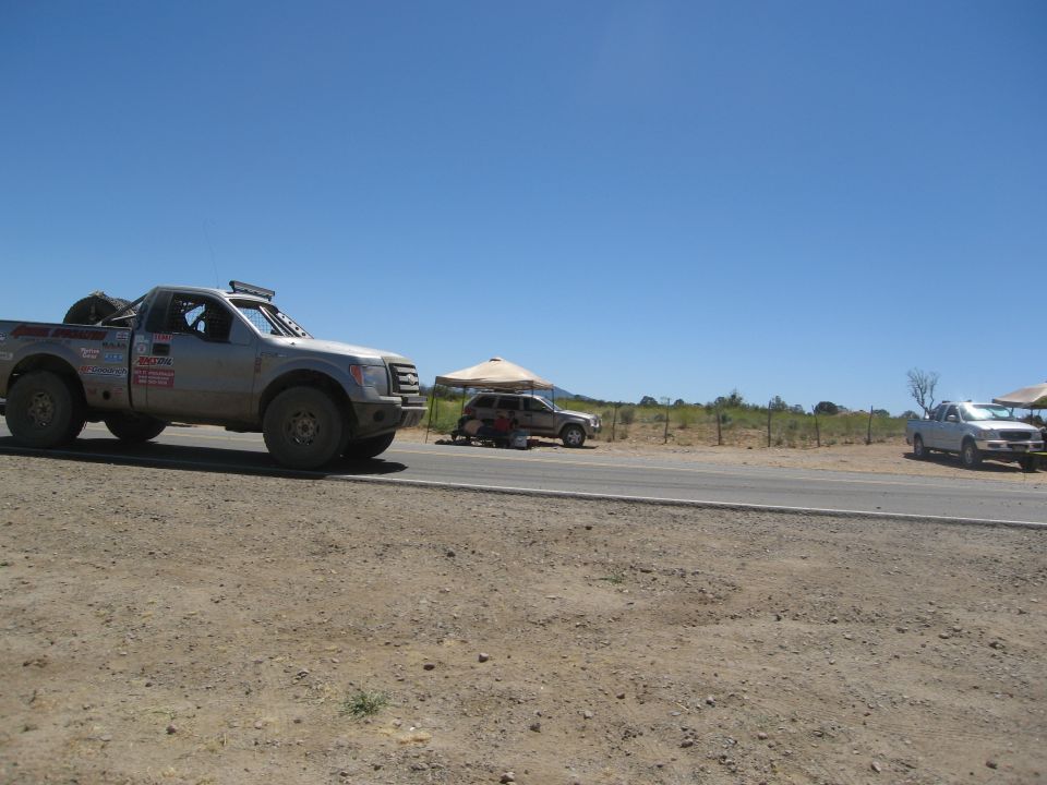 Silver pickup truck with off-road tires and light bar, parked on dirt road. Another SUV and truck visible in background