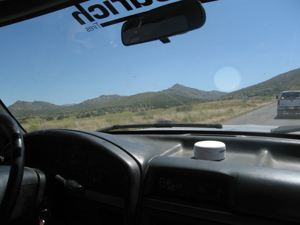 Dashboard interior with a white container on it, looking out the windshield at a mountain landscape and a white pickup truck on the road