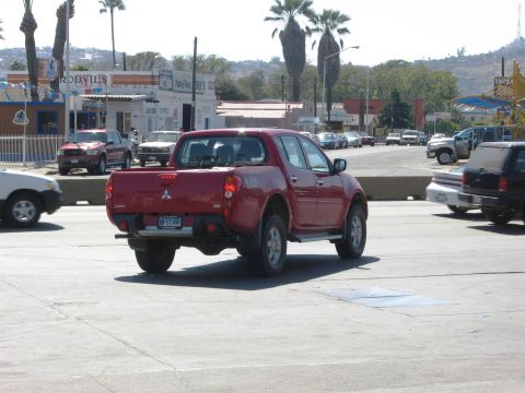 1986 Ford Bronco - A red pickup truck is seen from behind driving on a street with other vehicles and buildings in the background.
