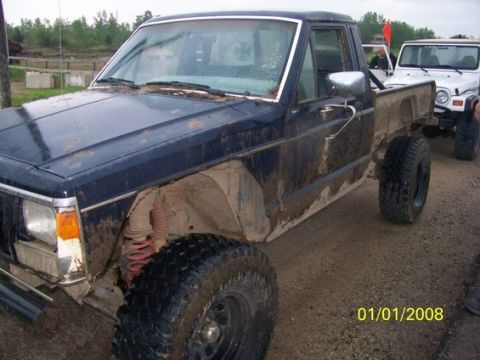 1987 Jeep Comanche - Two off-road Jeeps, one a dark blue pickup and the other a white Wrangler, are covered in mud.