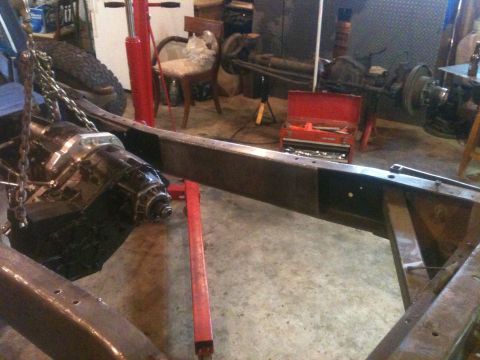 1976 Ford F-100 - Transmission suspended by an engine hoist over a bare truck frame in a workshop.