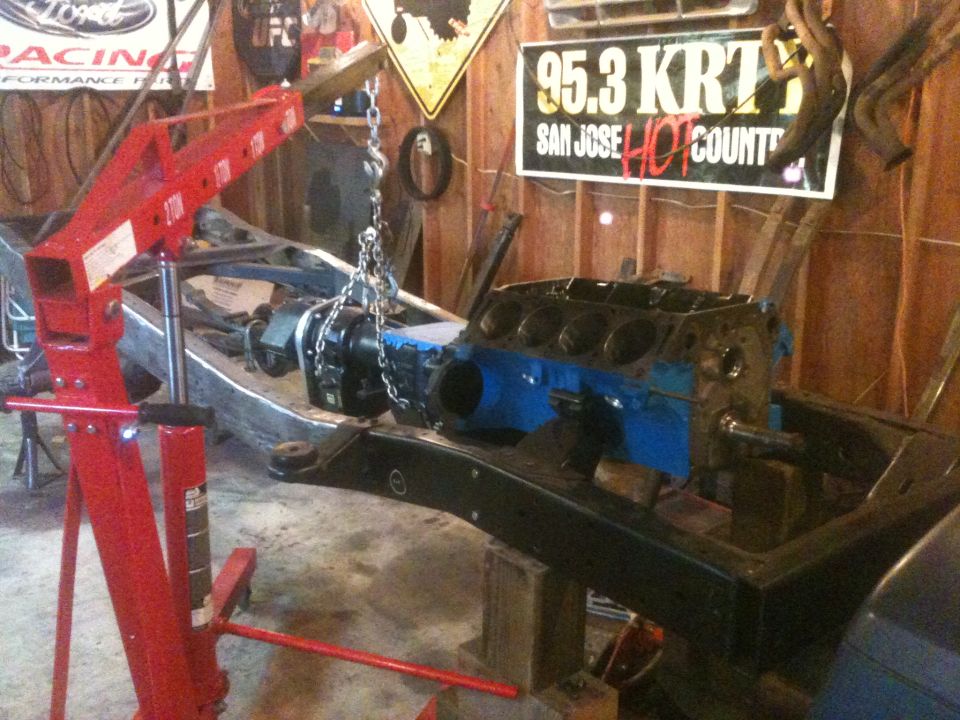 Engine block suspended by chains over a vehicle frame in a garage. Red engine hoist is visible