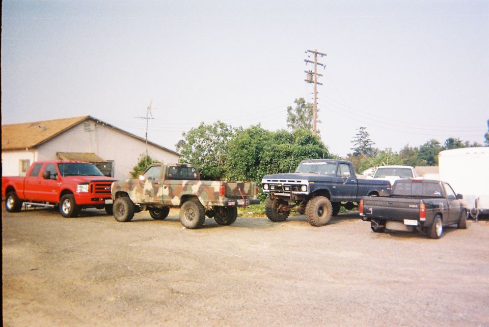 Four pickup trucks parked in a lot. A red Ford F-series, a camo-painted truck, a dark blue lifted truck, and a black pickup