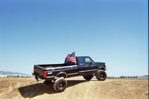 1992 Ford F-250 - A lifted 1992 Ford F-250 pickup truck flies an American flag while parked on a dirt incline under a clear blue sky.