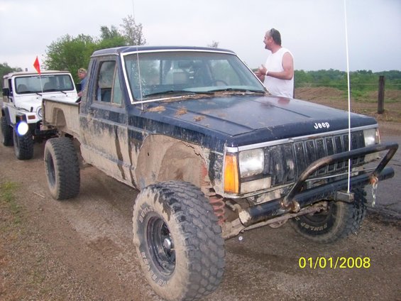Mud-covered 1987 Jeep Comanche pickup truck, dark blue, with a custom front bumper and large tires. Another Jeep is visible behind it