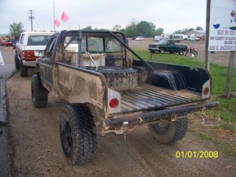 1987 Jeep Comanche - A heavily modified 1987 Jeep Comanche pickup truck is seen from behind, covered in mud, with a roll cage and spare tire visible.