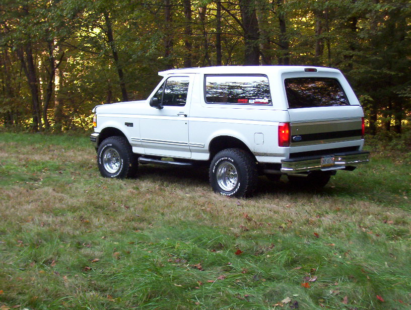 White lifted Ford Bronco SUV with BFGoodrich all-terrain tires parked on grass near woods, rear three-quarter view