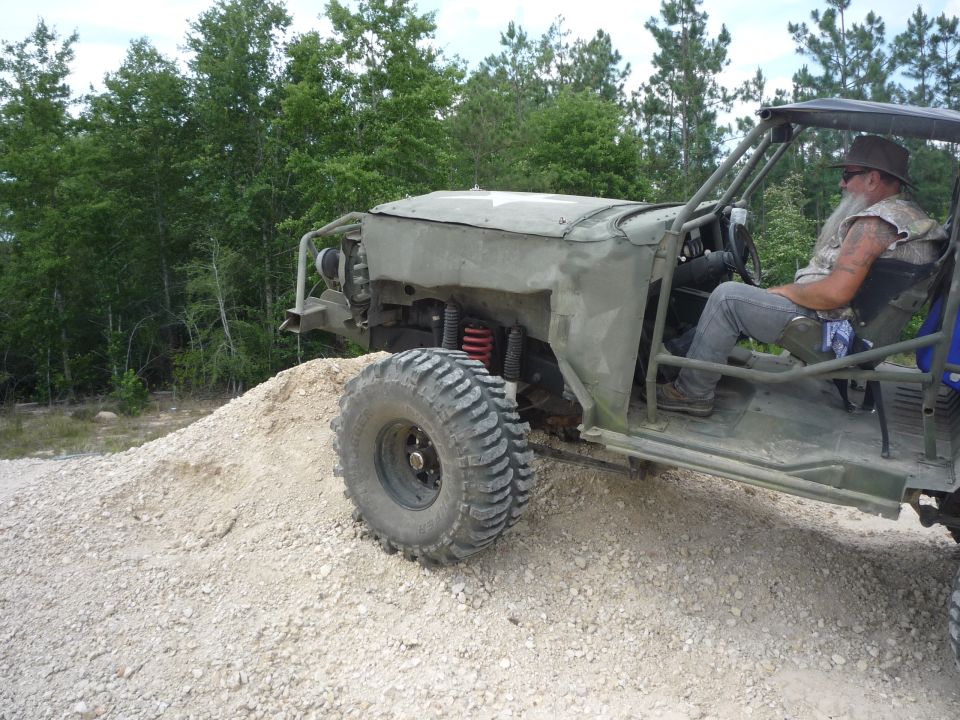 Custom off-road buggy with large tires, driven by a man wearing a hat and vest, on a dirt mound